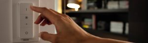 "Mão ajustando o dimmer LED inteligente Philips, controlando a iluminação ambiente em uma sala de estar aconchegante com estantes."