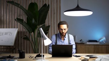 Um homem está sentado em uma mesa com um laptop à sua frente, iluminado por uma luminária suspensa e uma lâmpada para mesa à sua direita.