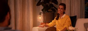 "Ambiente aconchegante de sala de estar criado por uma luminária de mesa LED de luz quente, iluminando uma mulher de suéter amarelo curtindo uma conversa noturna."